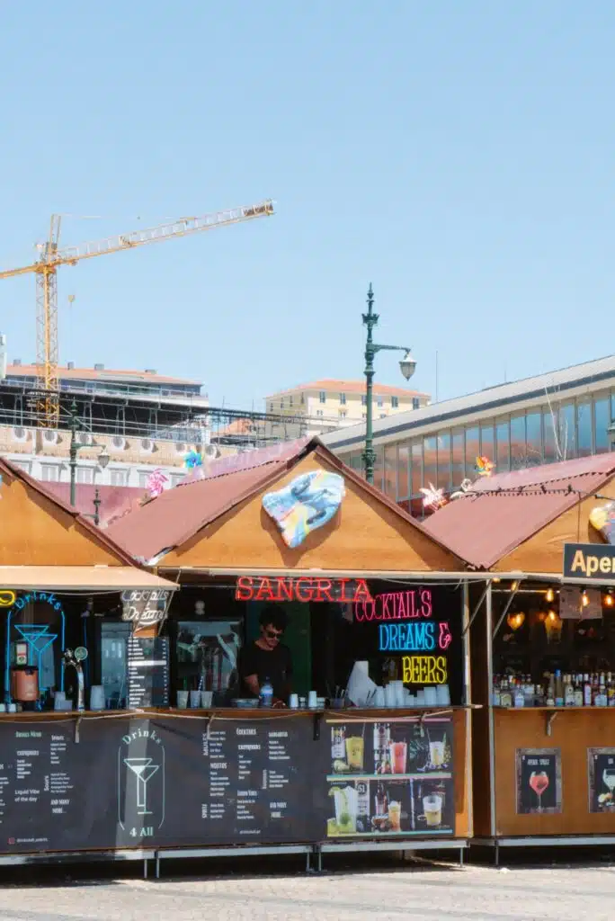 Wooden outdoor market stalls with colorful signs advertise sangria, cocktails, and beer—an essential stop during 3 days in Lisbon. A construction crane and modern buildings rise in the background under a clear blue sky.