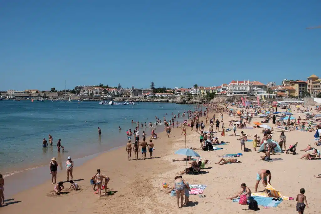 People relax, sunbathe, and swim on a sandy beach by calm blue water, with buildings and houses in the background. A perfect spot to visit during 3 days in Lisbon, the beach is busy with families and groups enjoying the sunny day.