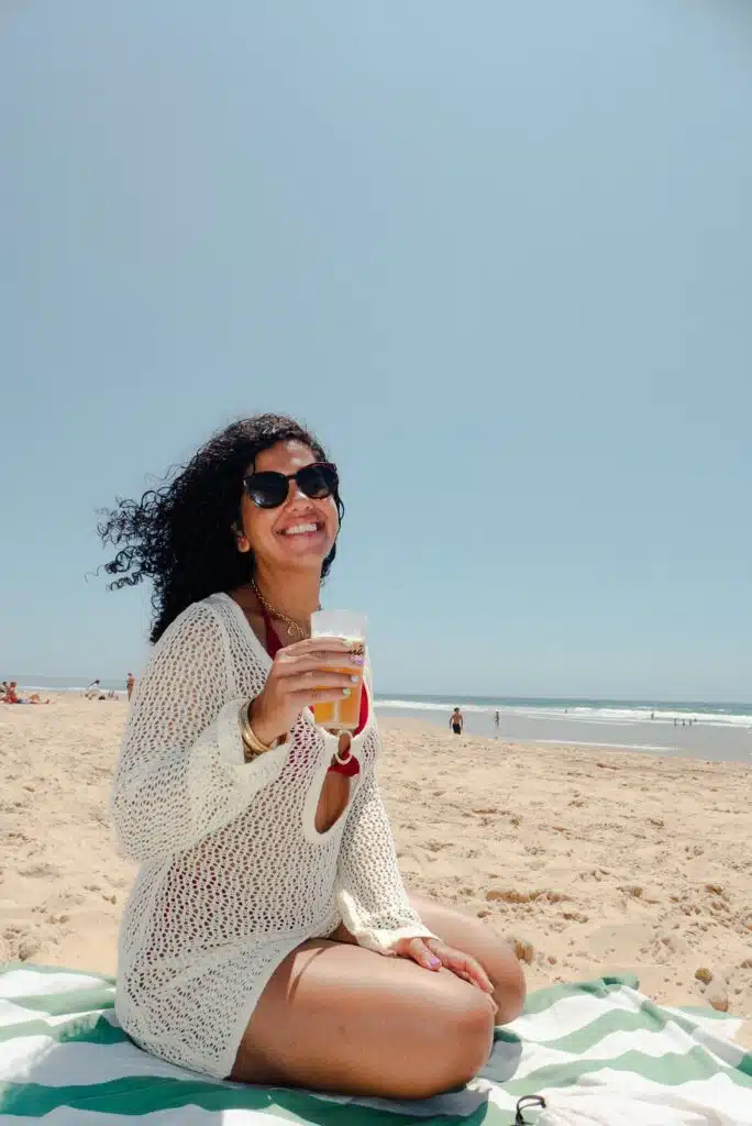 A woman wearing sunglasses and a white crochet beach cover-up sits on a striped towel at a sandy beach in Lisbon, smiling and holding a drink. The clear blue sky and ocean set the perfect scene for enjoying 3 days in Lisbon.