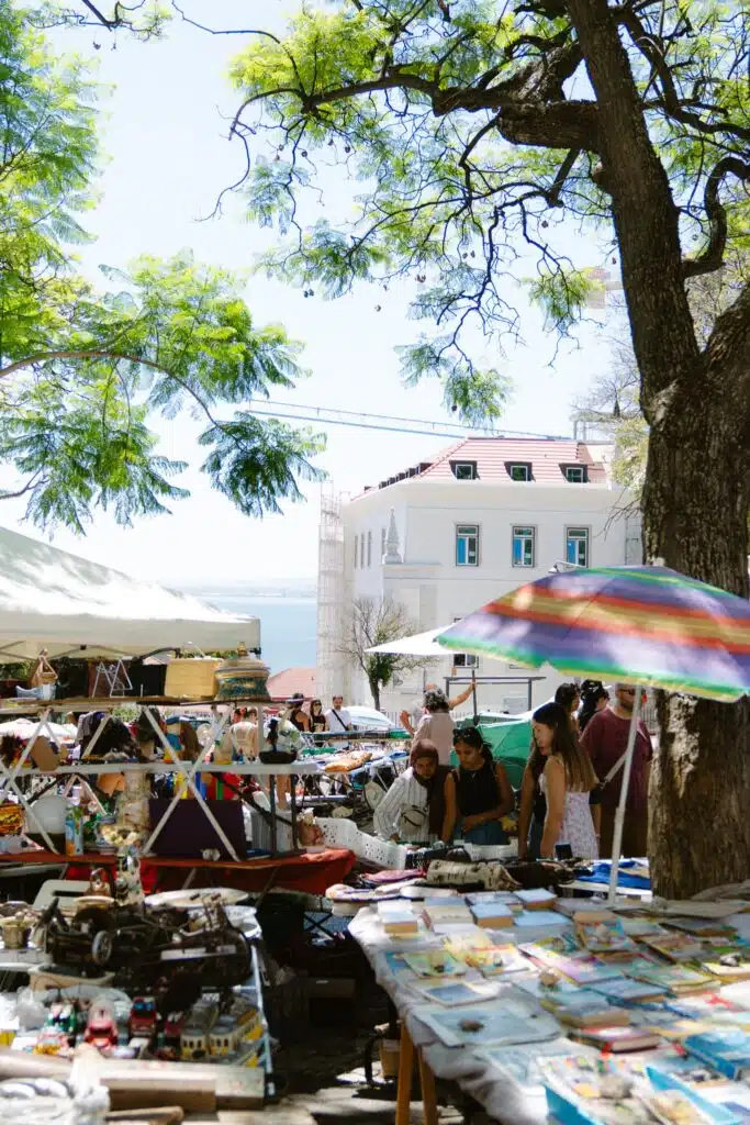 People browse colorful market stalls filled with various goods and antiques under large trees and a rainbow umbrella—a perfect scene to experience during 3 days in Lisbon, with a light-colored building and blue sky in the background.