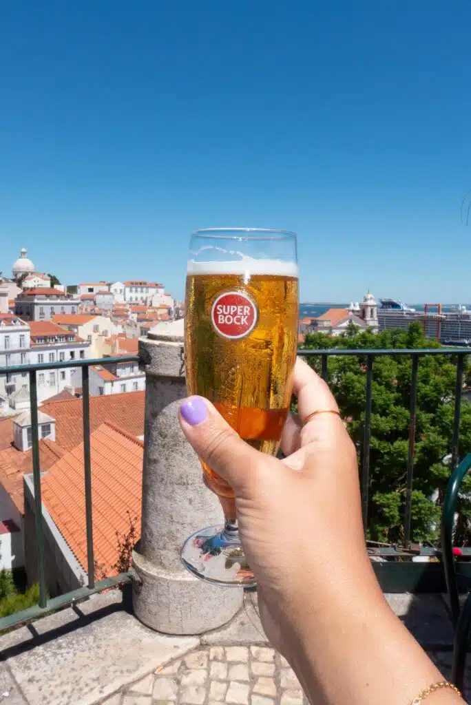 A hand with purple nail polish holds a glass of Super Bock beer; Lisbon's red rooftops and historic buildings stretch under a clear blue sky—the perfect view during 3 days in Lisbon.
