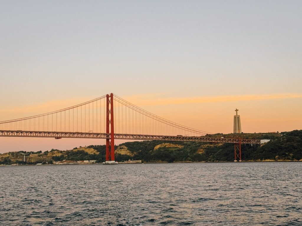 A red suspension bridge spans over a wide river at sunset, with a large statue of Christ atop a hill in the backgroundโan iconic view often admired during a Lisbon sunset cruise beneath softly glowing orange and pink skies.