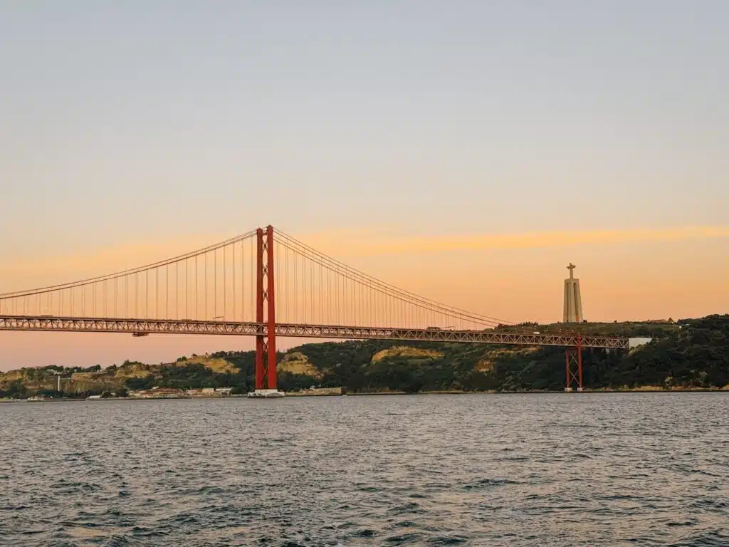 A red suspension bridge spans over a wide river at sunset, with a large statue of Christ atop a hill in the background—an iconic view often admired during a Lisbon sunset cruise beneath softly glowing orange and pink skies.