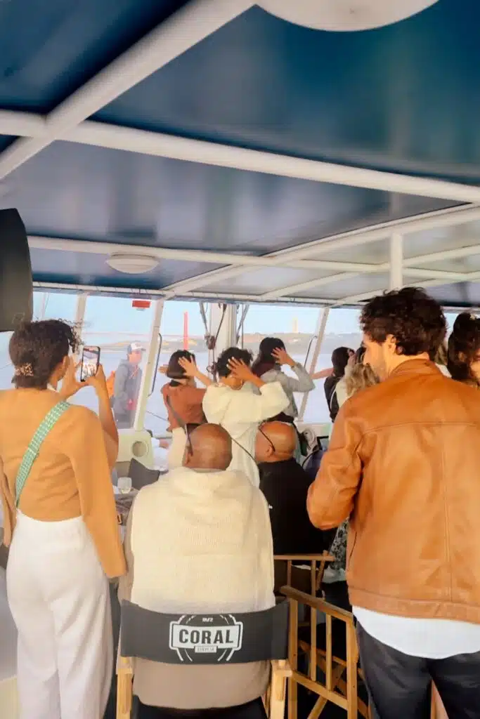 A group of people stand and sit on a boat’s covered deck during a sunset cruise in Lisbon, some taking photos and looking out at the water through large windows. One person wears a brown jacket, and another has a green strap bag.