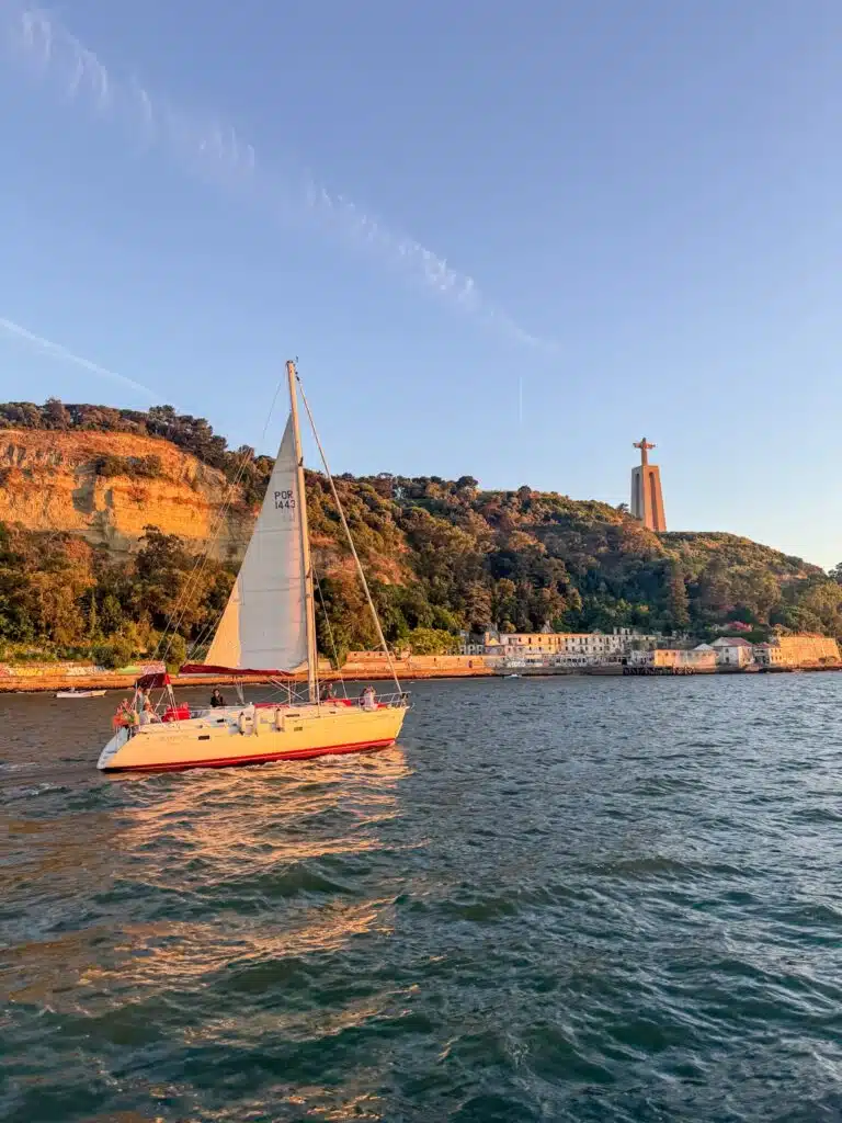 A white sailboat glides on a sunlit river near a hilly, green shore. In the background, the Christ statue stands atop a hill, glowing in golden sunlight—a perfect scene for a Lisbon sunset cruise.