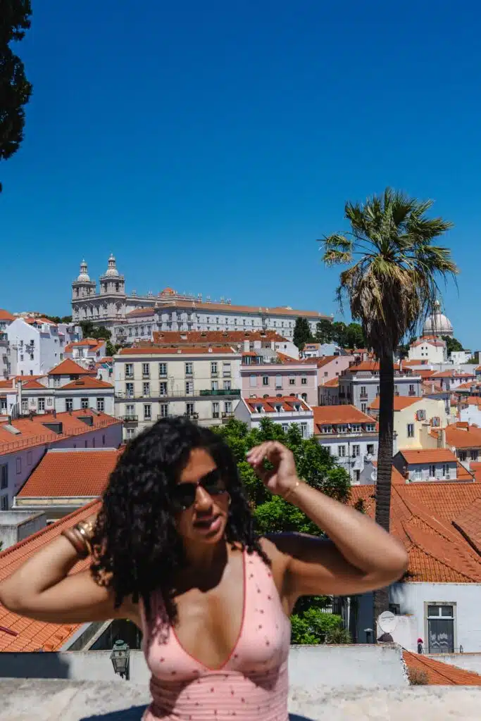 A woman with curly hair and sunglasses stands in the foreground, adjusting her hair, with a scenic view perfect for 3 days in Lisbon—red-roofed buildings, a palm tree, and a historic hilltop church beneath a clear blue sky.