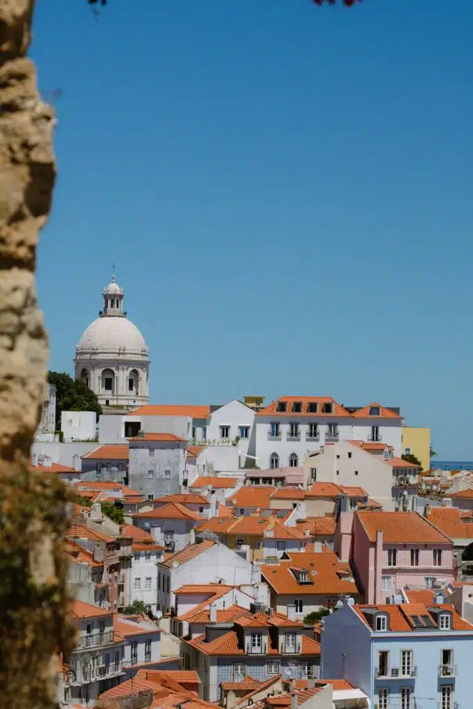 View of a cityscape with red-tiled rooftops and colorful buildings under a clear blue sky; a large domed church rises in the background, partially framed by a rough stone wall—perfect for capturing memories from 3 days in Lisbon.