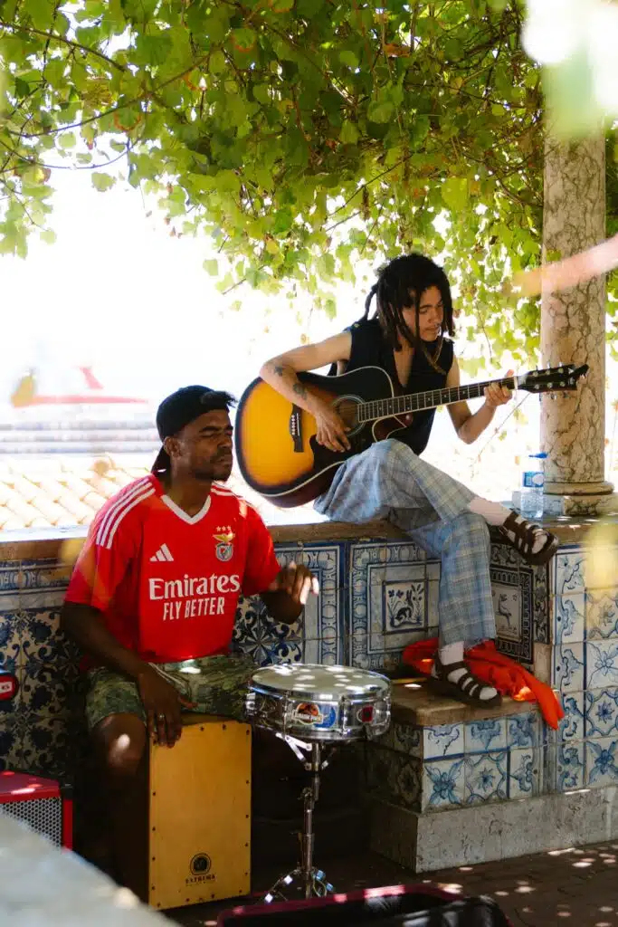 Two street musicians perform outdoors, offering a lively soundtrack for anyone spending 3 days in Lisbon. One plays a drum in a red football shirt, while the other, with dreadlocks, strums an acoustic guitar beneath sunlit, leafy branches.