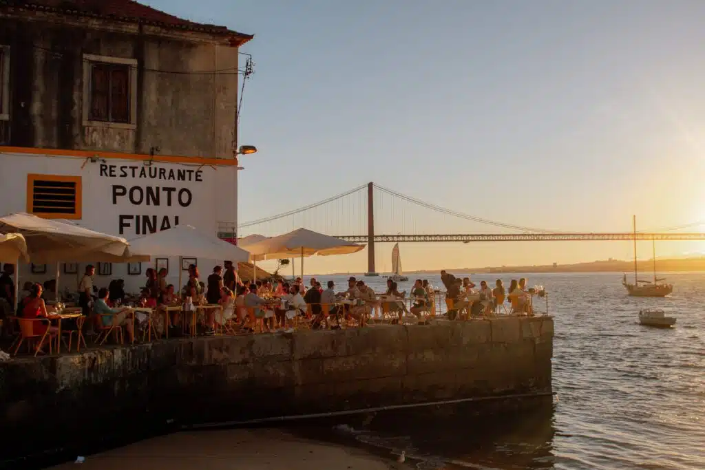 People dine outdoors at Restaurante Ponto Final, located on a riverside terrace at sunset. It's a must-stop during your 3 days in Lisbon, with the 25 de Abril Bridge and boats in the background creating a warm, scenic atmosphere.