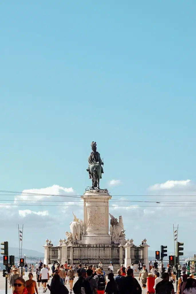 A large statue of a man on horseback stands in a busy city square under a clear blue sky, surrounded by people walking and traffic lights—a classic sight if you spend 3 days in Lisbon.