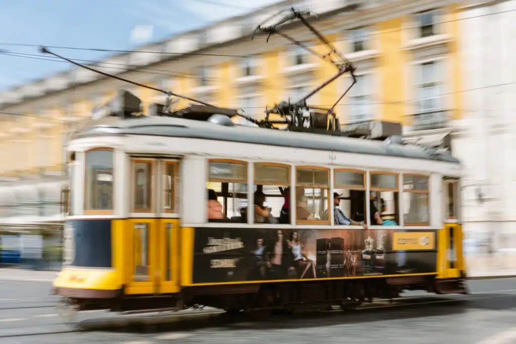 A yellow and white vintage tram moves quickly through a city square in Lisbon, with blurred buildings and people in the background—a classic scene to witness during 3 days in Lisbon.