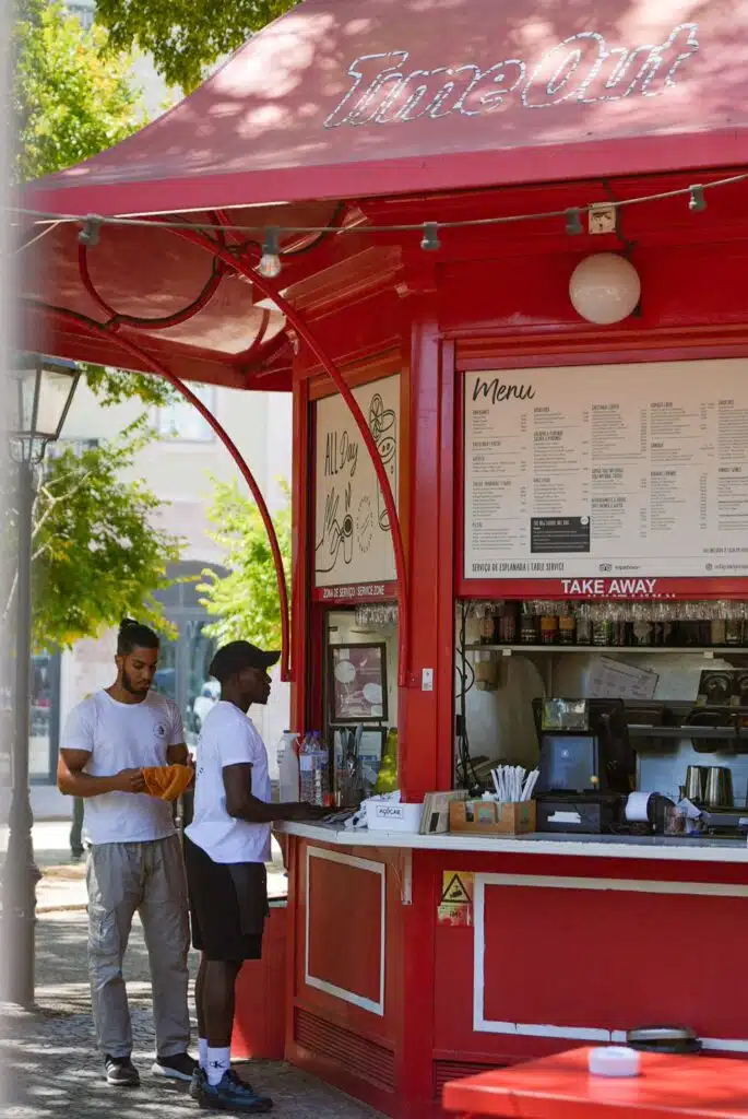 Two men stand at a bright red outdoor food kiosk called "TimeOut," reading the menu and placing an order on a sunny day—a perfect stop during 3 days in Lisbon, with green trees in the background.