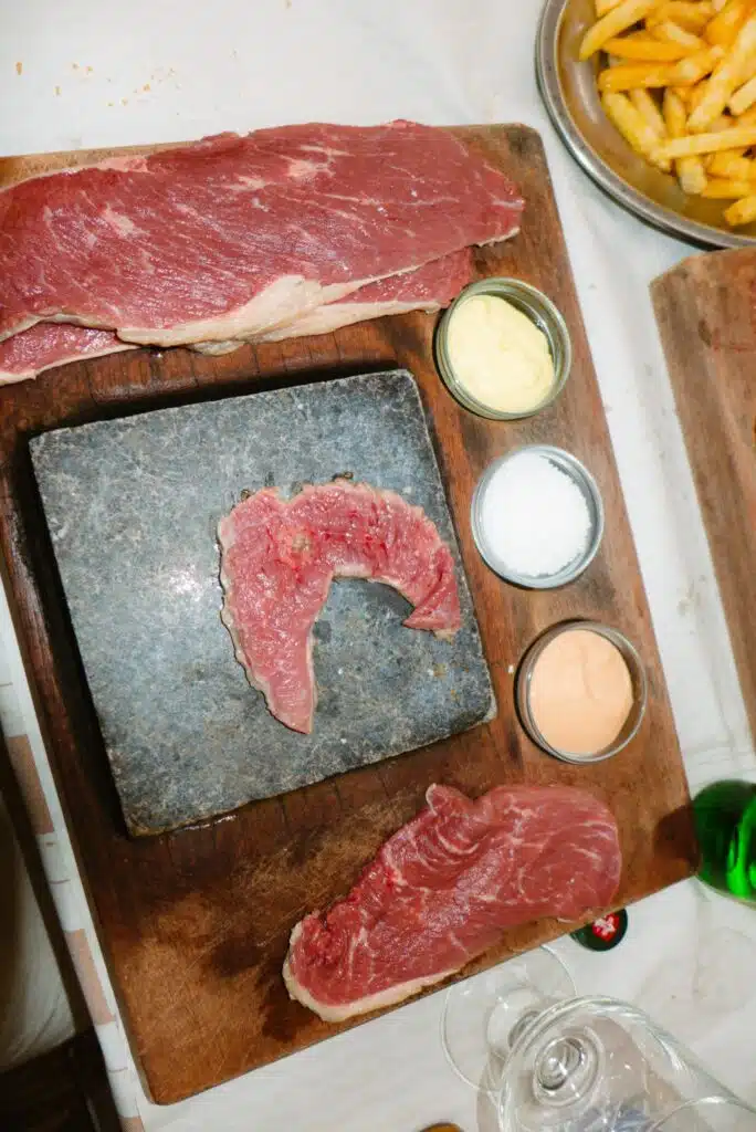 Raw steaks on a wooden board surround a hot stone, with three small jars of sauce and salt nearby; fries in a metal bowl complete this tempting scene—perfect after exploring 3 days in Lisbon’s vibrant food culture.