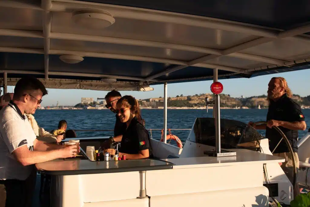 People on a boat enjoying drinks at a bar area, with a man steering the boat—a perfect sunset scene after 3 days in Lisbon, with warm sunlight gleaming over the water and a distant shoreline in the background.