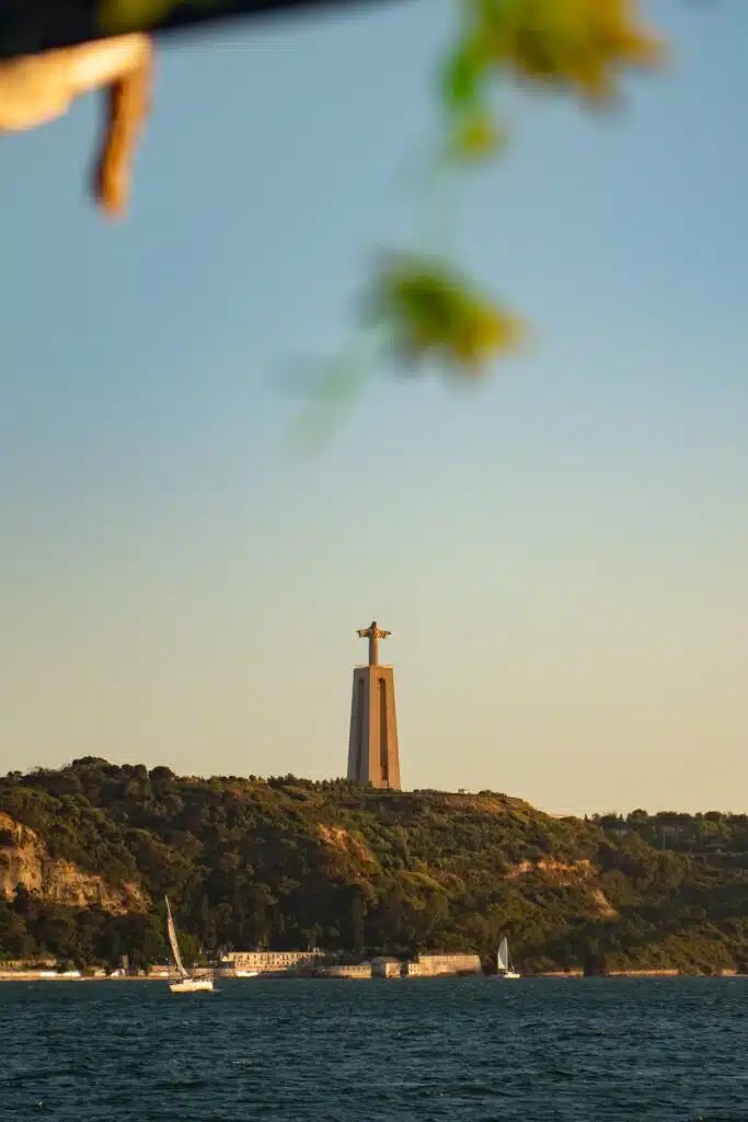 A tall statue of Christ with outstretched arms stands on a pedestal atop a green hill overlooking the water, under a clear sky—a must-see if you spend 3 days in Lisbon. Sailboats drift gracefully on the sparkling water in the foreground.