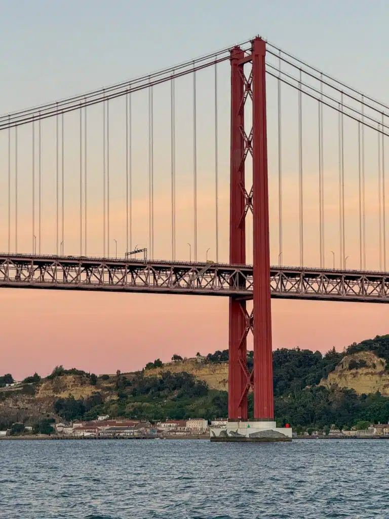 A tall red suspension bridge spans over a body of water at sunset, with hills and buildings on the far shore beneath a pastel pink and blue sky—an unforgettable sight during 3 days in Lisbon.