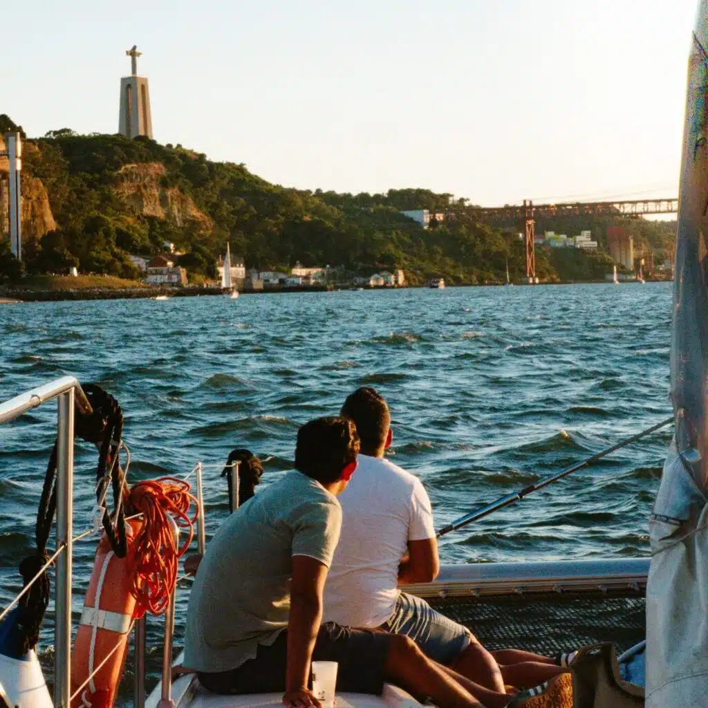 Two people sit on a boat facing the water as the sun sets over Lisbon’s hilly coastline, with the iconic Christ statue and a large bridge in view—an unforgettable moment during 3 days in Lisbon.