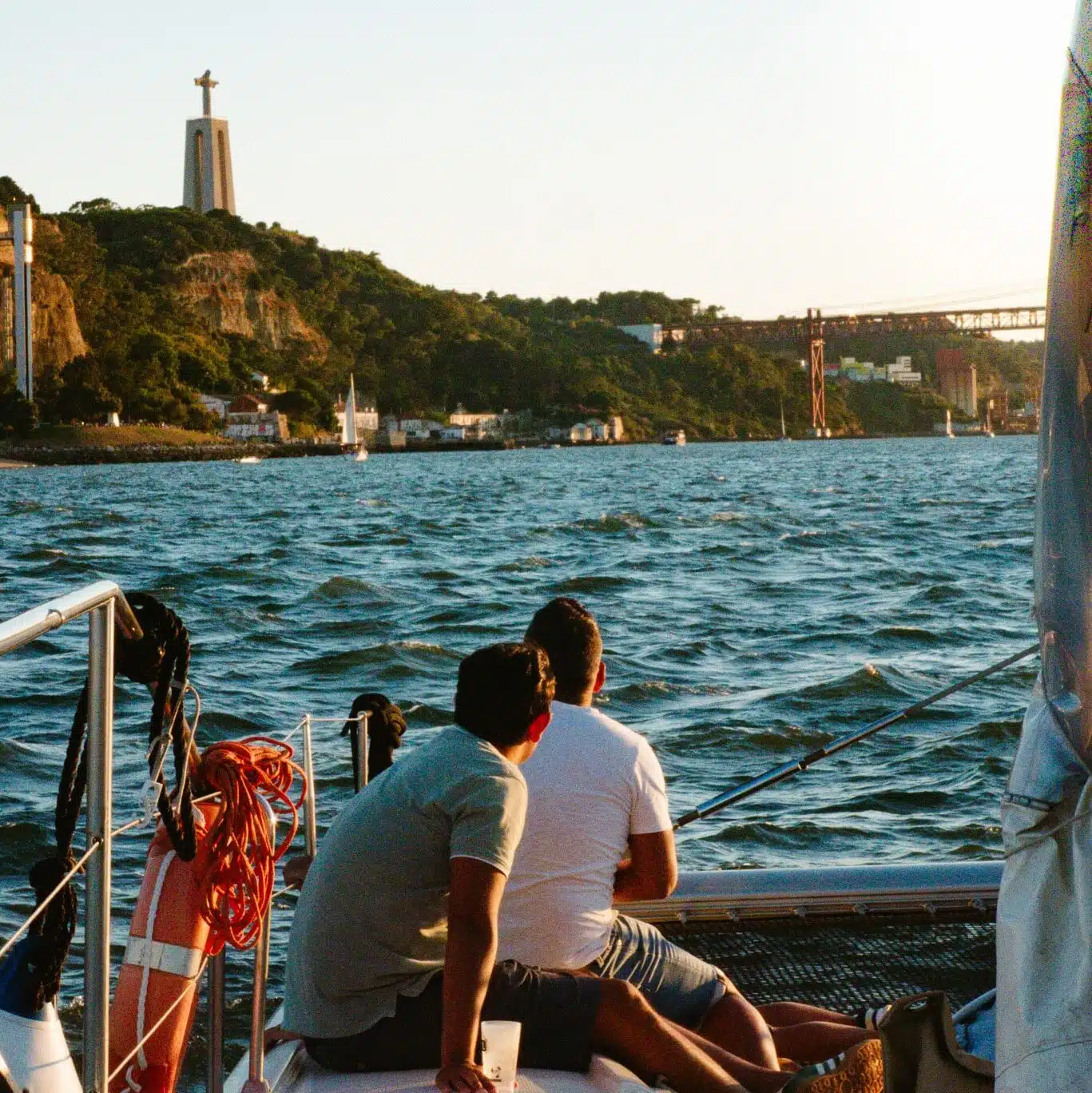 Two people sit on a boat facing the water as the sun sets over Lisbon’s hilly coastline, with the iconic Christ statue and a large bridge in view—an unforgettable moment during 3 days in Lisbon.