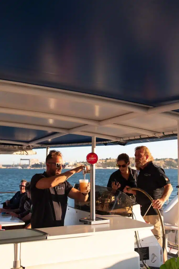 A man pours a beer at an outdoor bar on a boat, while two others stand nearby and others sit in the background—capturing the perfect moment for those enjoying 3 days in Lisbon with stunning water and shoreline views in the sunlight.