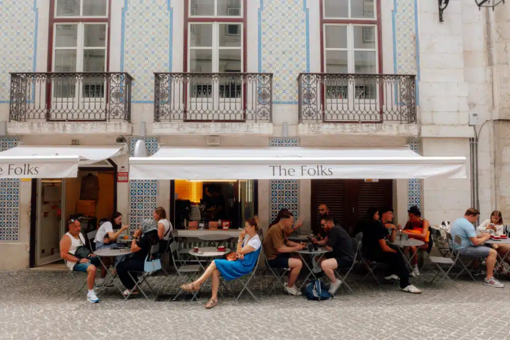 People sit at outdoor tables under a white awning labeled "The Folks," in front of a building with patterned tiles and tall windows—a charming café scene perfect for your 3 days in Lisbon, with relaxed diners enjoying the lively cobblestone street.