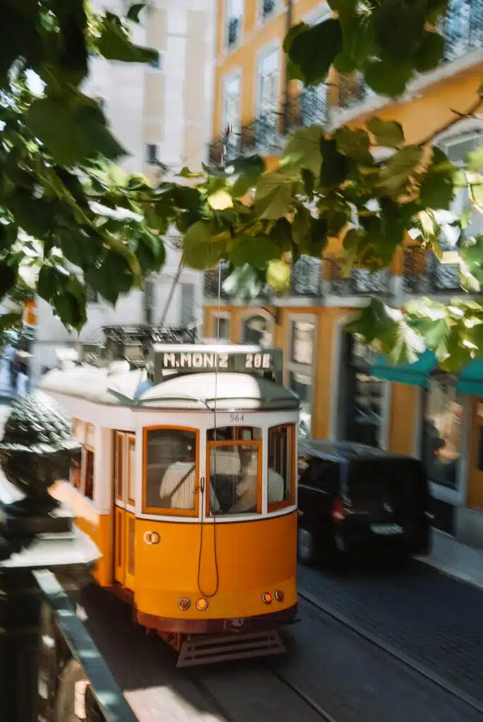 A yellow tram travels through a narrow, sunlit street lined with yellow buildings and leafy trees in Lisbon, Portugal. Perfect for exploring during 3 days in Lisbon, the tram’s destination sign reads "M.MONIZ 28E.