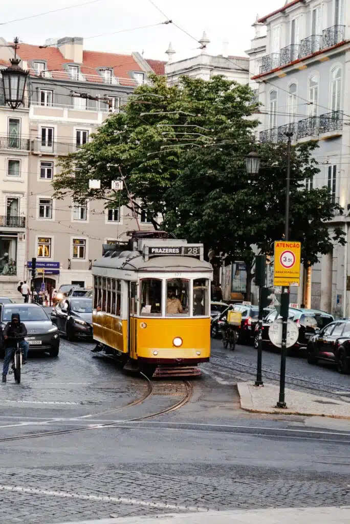 A yellow tram travels down a cobblestone street in Lisbon, perfect for exploring during 3 days in Lisbon, surrounded by parked cars, historic buildings, and trees on a cloudy day.