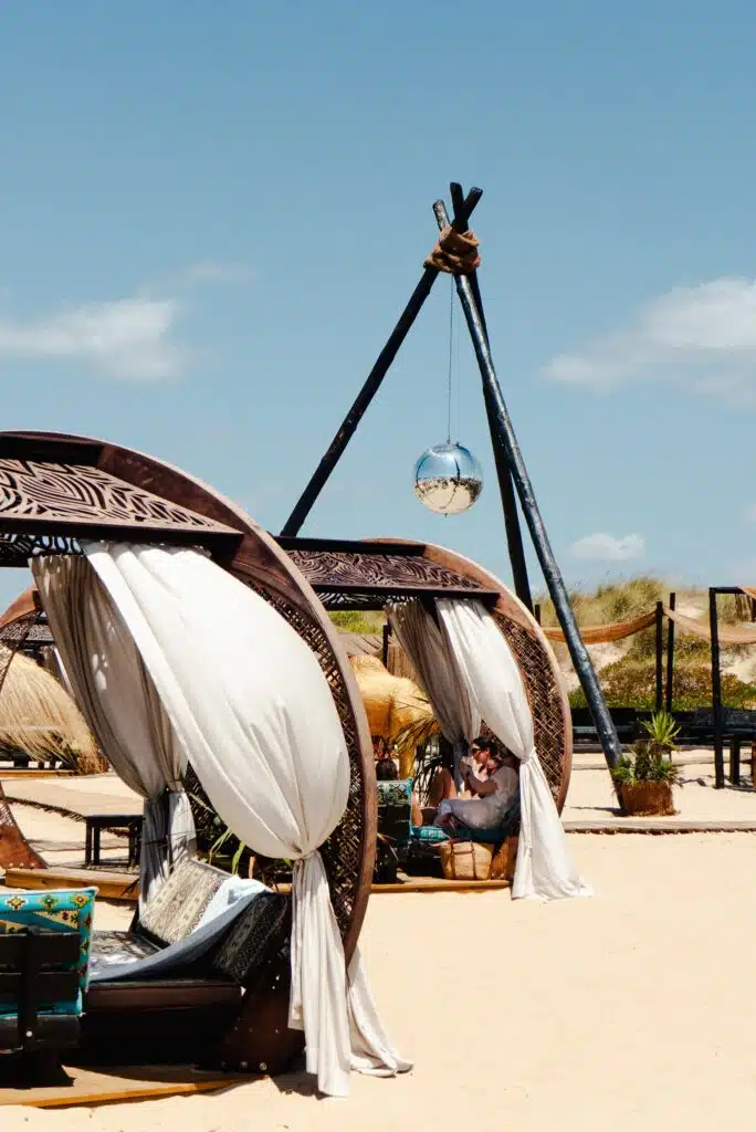 Two ornate wooden cabanas with white curtains stand on a sandy beach, offering a scene as dreamy as spending 3 days in Lisbon. A large tripod holds a reflective disco ball, with blue sky and dune grasses in the background.