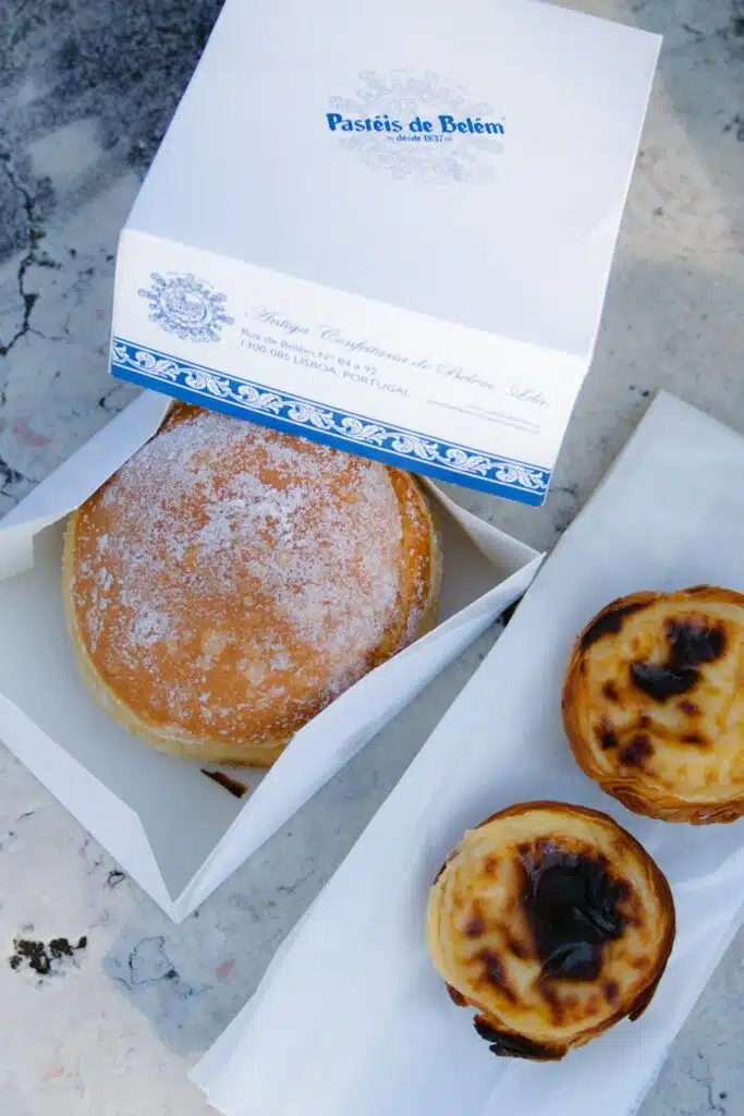 A white pastry box labeled “Pastéis de Belém” holds a sugar-coated round doughnut and sits next to two Portuguese custard tarts (pastéis de nata)—a delicious find for anyone spending 3 days in Lisbon—displayed on white paper atop a marble surface.
