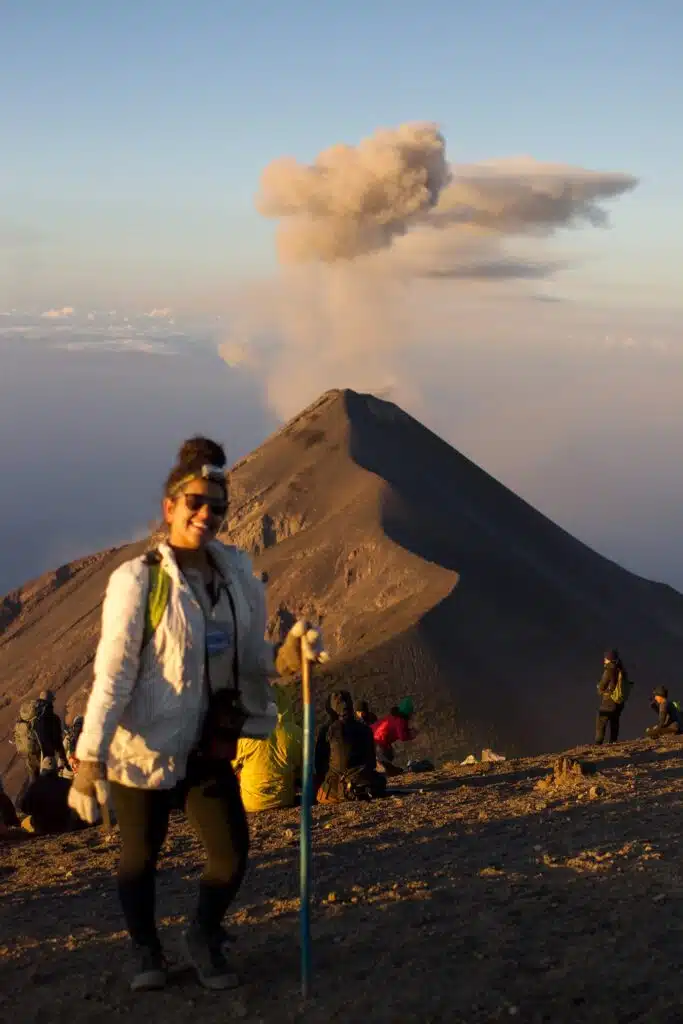 A person in hiking gear stands with a walking stick in the foreground, smiling on a mountain slope—a perfect setting for First-Time Solo Travel Destinations. In the background, a volcano emits ash as people sit and observe the scene.