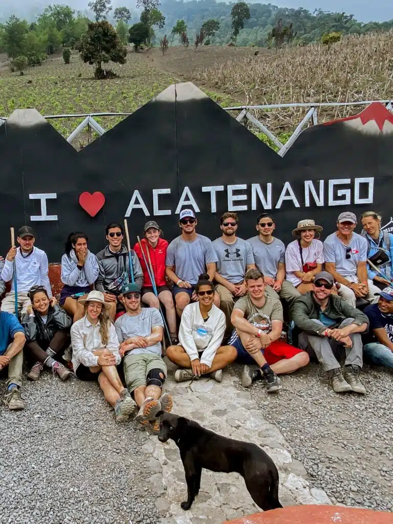 A group of people poses in front of a sign that reads “I ♥ ACATENANGO,” with mountains and fields behind them. A black dog stands in the foreground. Acatenango is one of the top First-Time Solo Travel Destinations for adventurers.
