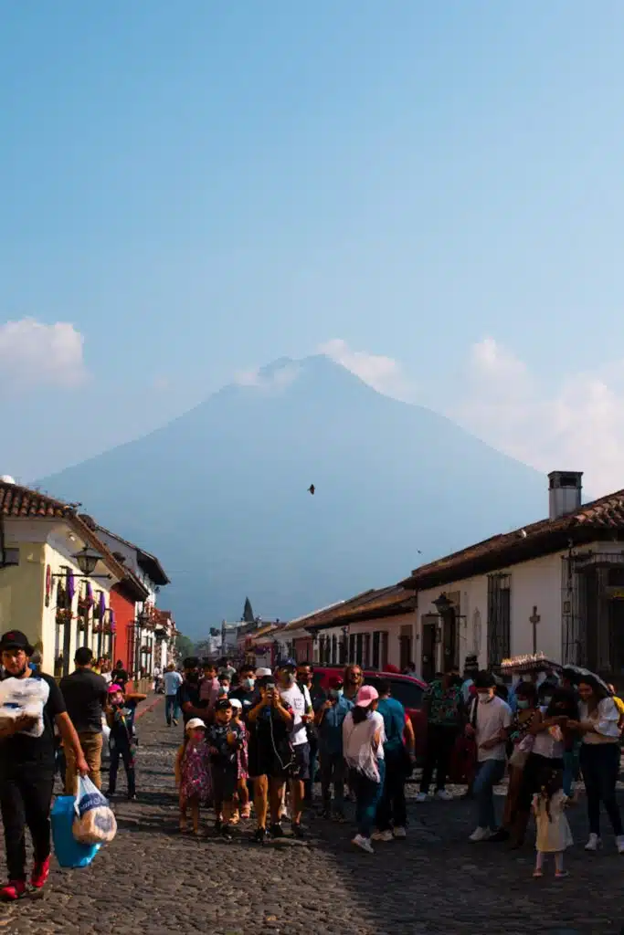 A crowd of people walks along a cobblestone street lined with buildings, with a large volcano and cloudy sky in the background—a picturesque scene perfect for those seeking First-Time Solo Travel Destinations.