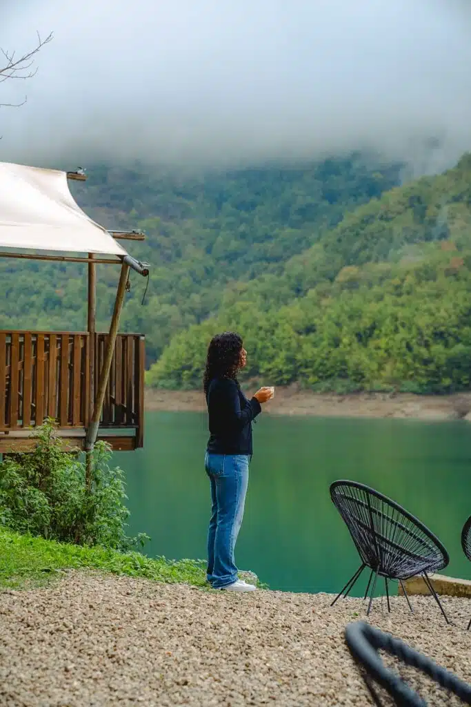 A person with curly hair stands holding a cup, facing a calm lake surrounded by misty, forested hills—an ideal setting for those seeking First-Time Solo Travel Destinations. Nearby are black outdoor chairs and a wooden cabin with a canopy.