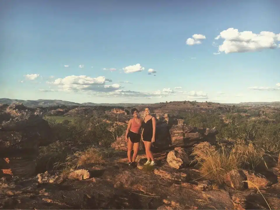 Two women stand on rocky terrain surrounded by grass and shrubbery, framed by distant hills under a partly cloudy sky. The warm sunlight highlights this inviting scene—perfect inspiration for First-Time Solo Travel Destinations.