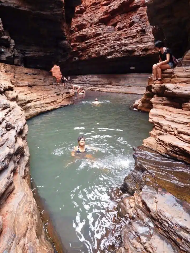 People swim in a natural pool surrounded by layered red and brown rock walls in a gorge—a perfect spot for first-time solo travel destinations. One person sits on a rock ledge, while others enjoy the water in this scenic, rocky landscape.