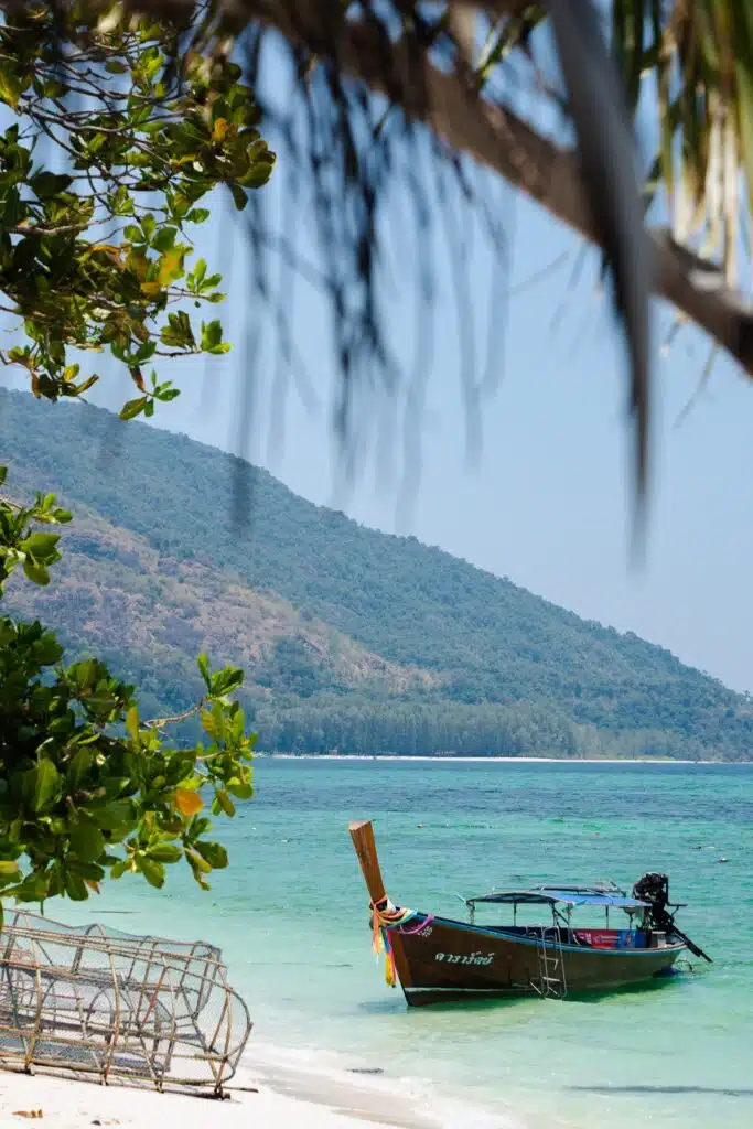 A wooden long-tail boat floats on clear turquoise water near a sandy beach, with green trees and fishing cages in the foreground—an inviting scene for first-time solo travel destinations—set against a forested hill beneath a blue sky.