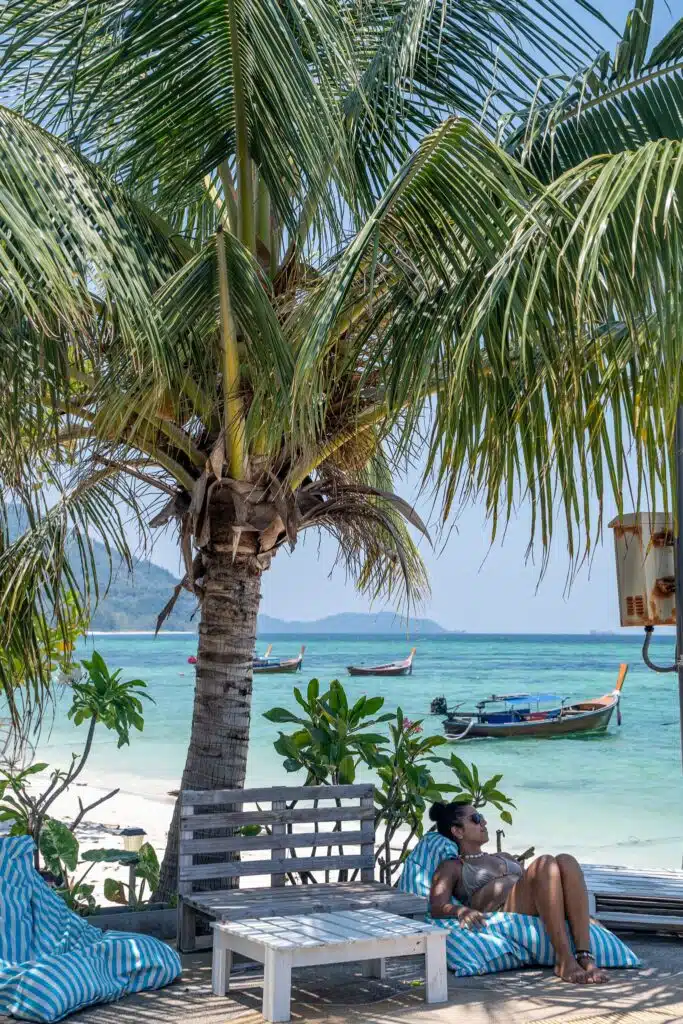 A woman relaxes on a lounge chair under a palm tree by the beach, with blue and white cushions, clear turquoise water, and traditional boats—an idyllic scene perfect for First-Time Solo Travel Destinations.