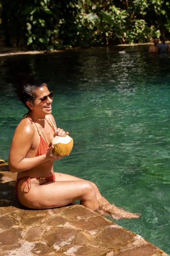 A woman in a red bikini sits on the edge of a stone pool, smiling and holding a coconut drink—capturing the relaxed vibe of First-Time Solo Travel Destinations—with green water, lush plants, and sunlight reflections in the background.