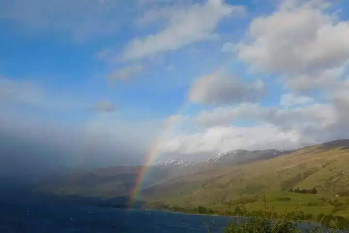 A faint rainbow arcs across a partly cloudy sky over blue water, green hills, and distant snow-capped mountains—an inviting scene for those seeking picturesque First-Time Solo Travel Destinations.