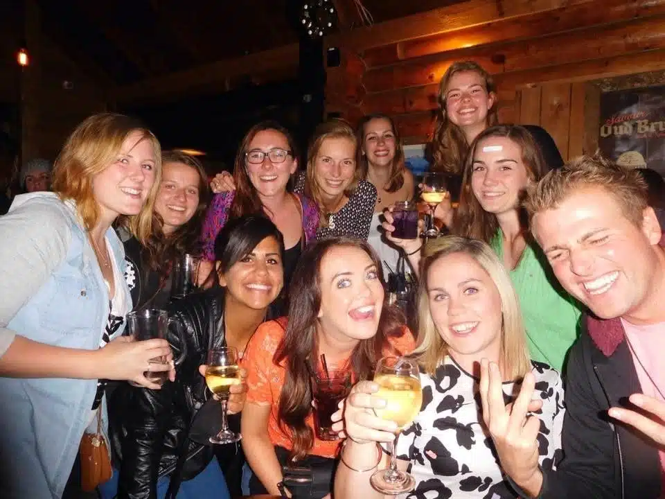 A group of smiling friends, mostly women, pose together indoors at night, celebrating and sharing stories about First-Time Solo Travel Destinations. They appear happy and cheerful, with wooden walls and warm lighting in the background.