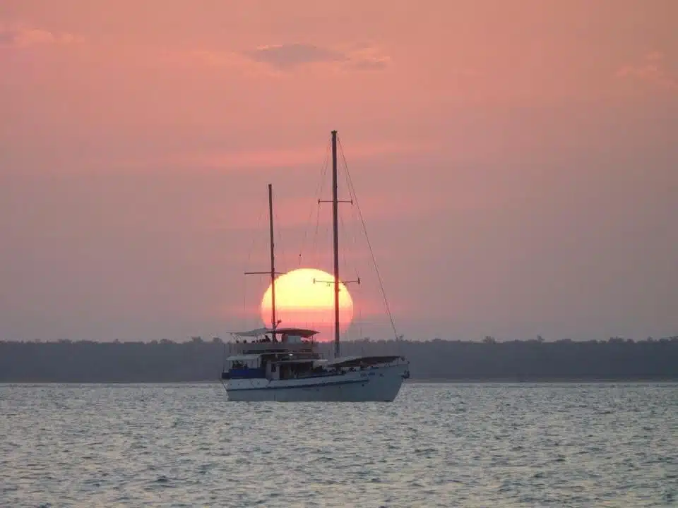 A sailboat floats on calm water at sunset, with the large orange sun low in the sky behind the boat and a tree-lined shore in the background—an inspiring scene for those seeking first-time solo travel destinations.