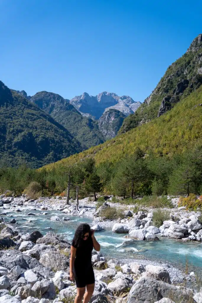 A woman in a black dress stands on rocky terrain near a clear blue river, with green forested mountains beyond—a perfect scene for those seeking First-Time Solo Travel Destinations under a bright blue sky.