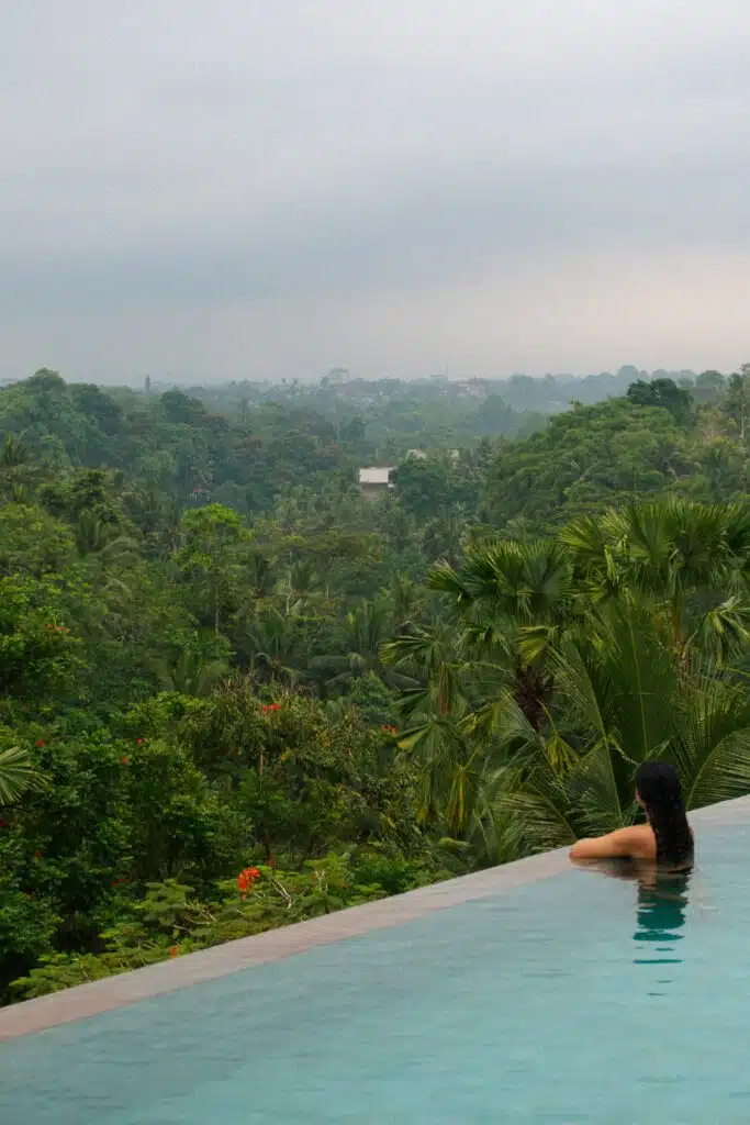 A person with long dark hair relaxes in an infinity pool overlooking a lush, green tropical forest under a cloudy sky—capturing the serenity found at some of the best First-Time Solo Travel Destinations.