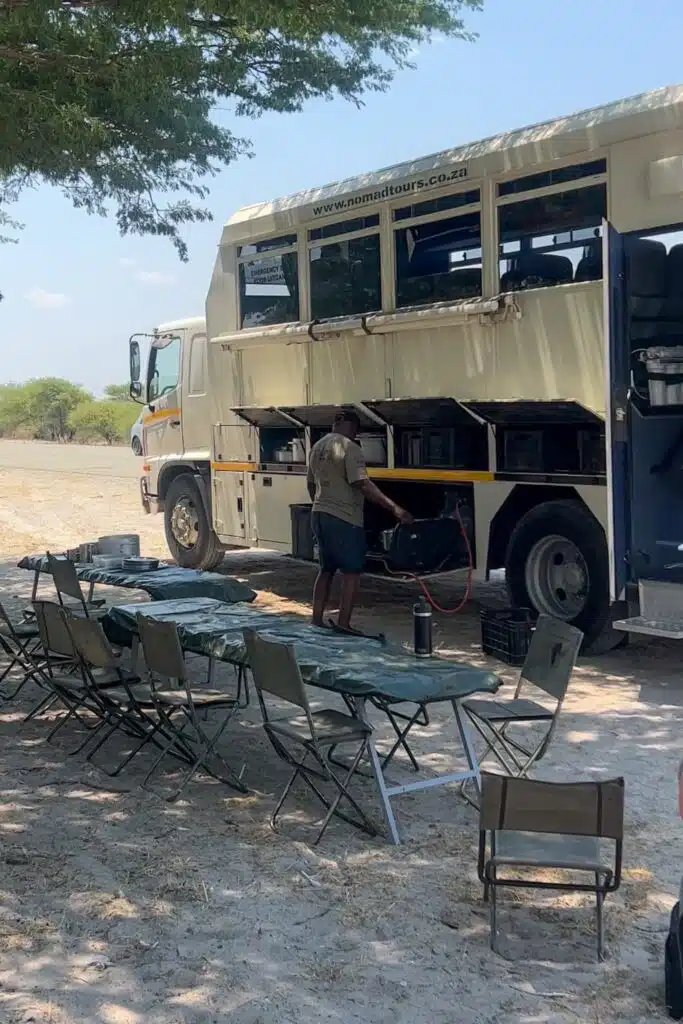 An African Overland Tours bus with open compartments is parked beside a table and several camping chairs set up under a tree. A person stands near the bus, preparing equipment on this sunny outdoor day.