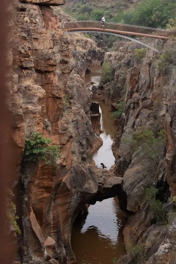 A narrow, rocky canyon with reddish-brown cliffs and a muddy river flowing through it, perfect for African Overland Tours. A natural rock arch arches over the water while a footbridge above lets people enjoy the sparse greenery on the rocks.