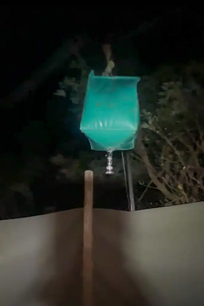 A green plastic bag filled with water hangs above a wall at night, reminiscent of makeshift showers seen on African Overland Tours, with trees in the background. A tap is attached to the bottom, and a wooden stick leans against the wall below.
