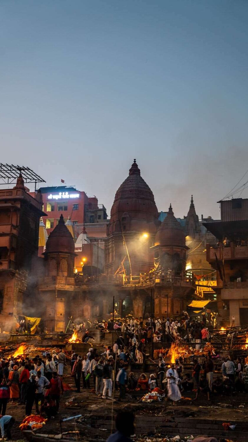 A bustling scene at a ghats during twilight, with numerous people gathered around multiple burning pyres. The backdrop features traditional domed structures and buildings, illuminated by streetlights, creating a spiritual and historic atmosphere - Marnikanika Ghat Varanasi