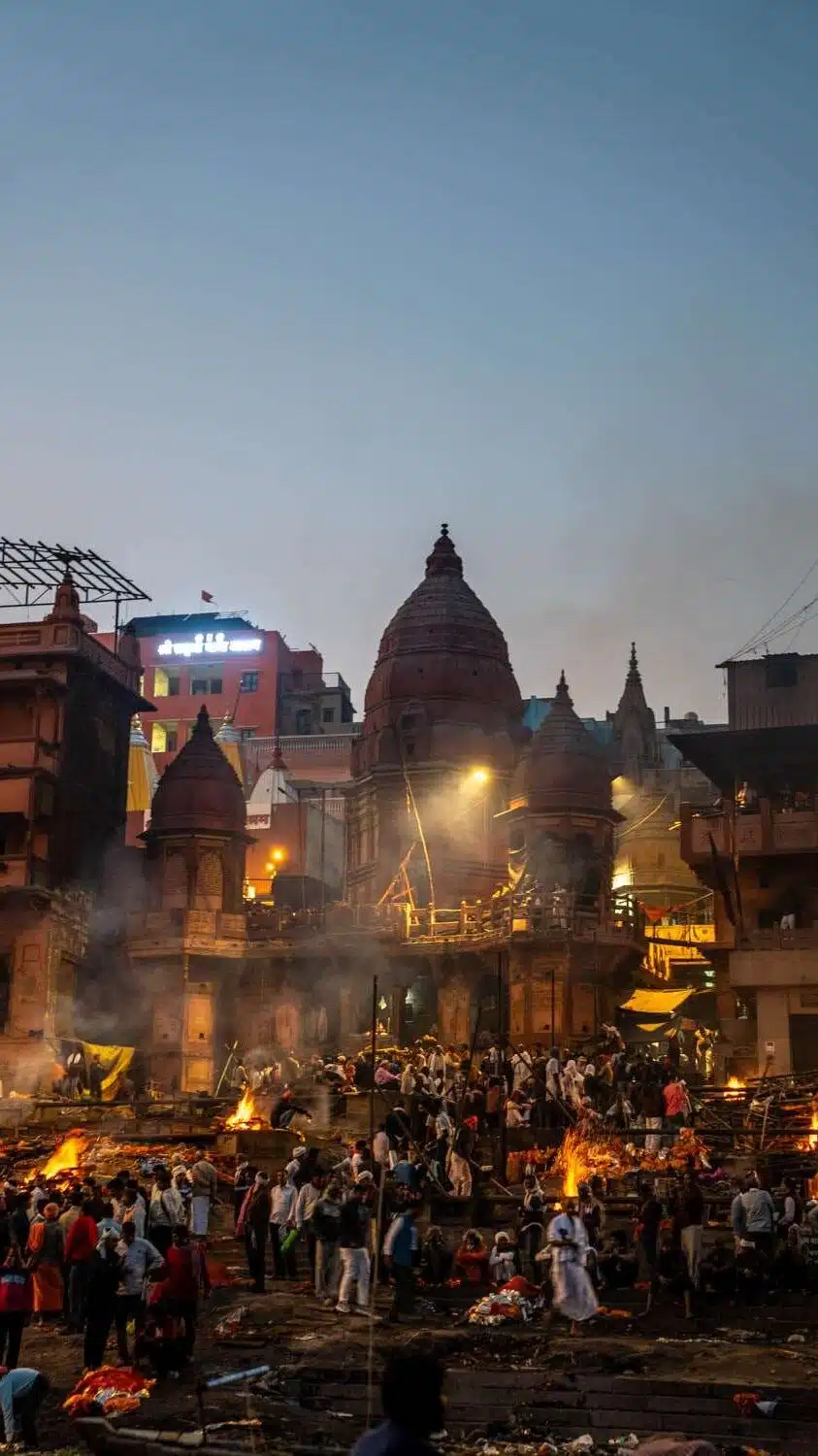 A bustling scene at a ghats during twilight, with numerous people gathered around multiple burning pyres. The backdrop features traditional domed structures and buildings, illuminated by streetlights, creating a spiritual and historic atmosphere - Marnikanika Ghat Varanasi