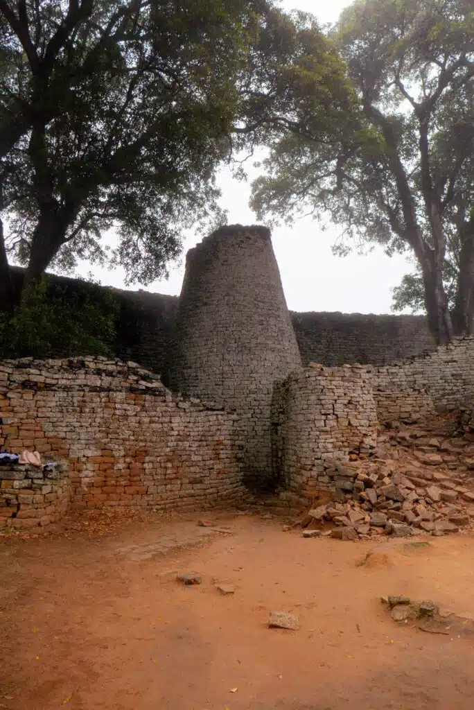 Ancient stone ruins with a tall, conical tower rise amid trees and partial walls on a dirt ground, scattered with stones. Under an overcast sky, this site evokes the spirit of African Overland Tours and age-old adventures.