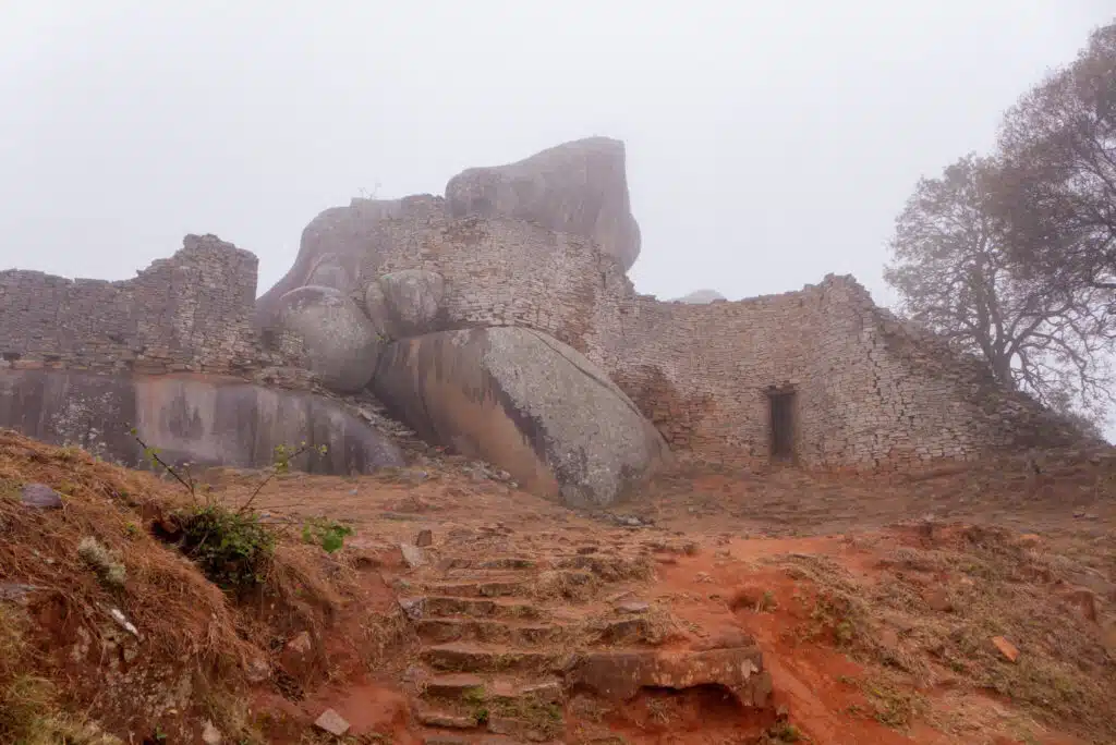 Ancient stone ruins partially surrounded by large boulders on a foggy day, with worn stone steps leading up to the structure and sparse vegetation on reddish-brown soil—an evocative scene often explored on African Overland Tours.