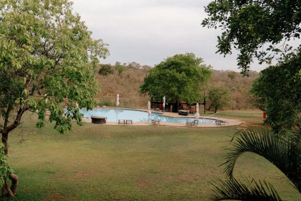 A circular outdoor swimming pool is surrounded by green grass, trees, lounge chairs, and umbrellas—offering a serene retreat for guests on African Overland Tours beneath a cloudy sky in a spacious, natural landscape.